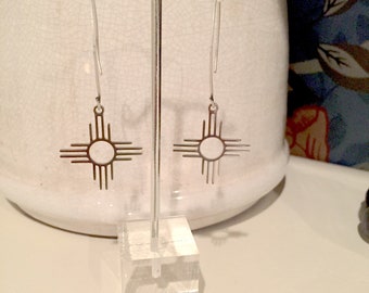 Könnte beinhalten: Ein Paar silberne Ohrringe mit einem Sonnensymbol-Design. Die Ohrringe hängen an einem durchsichtigen Acryl-Ständer.