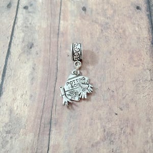 May include: A silver-colored charm with a gardening theme. The charm features a small bag of potting soil with the words "Potting Soil" and a pair of gardening gloves. The charm has a decorative top with a loop for attaching to a bracelet or necklace.