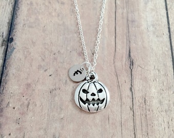 Op de afbeelding: Een zilveren ketting met een kleine pompoenbedel en een zilveren initialbedel. De pompoenbedel heeft een gesneden jack-o'-lantern gezicht.