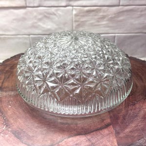 May include: Clear glass bowl with a textured, starburst pattern. The bowl has a rounded, dome-like shape and sits on a wooden surface. The rim of the bowl has a vertical ribbed design. The background is a white brick wall.