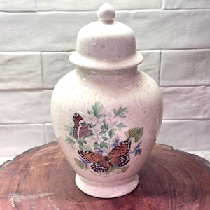 May include: A cream-colored ceramic jar with a lid, adorned with butterfly and floral designs. The jar has a speckled finish and a rounded shape. The butterflies are brown, orange, and black, with white and blue flowers.