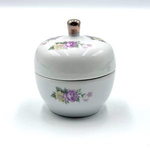 May include: A white ceramic lidded jar with a rounded shape, featuring a metallic gold-tone knob handle on top. The exterior is decorated with small floral clusters in shades of purple, yellow, and green on both the lid and the main body.