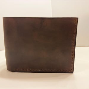 Puede incluir: Cartera de cuero marrón con costuras visibles en el borde inferior. La cartera tiene forma rectangular y una superficie ligeramente texturizada. El color es un marrón oscuro intenso, lo que sugiere un producto de cuero natural.