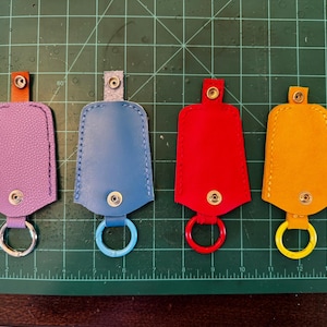 May include: Four leather keyrings in purple, blue, red, and yellow. Each has a matching ring and a snap closure. The keyrings are displayed on a green cutting mat. Measurements are in centimetres.