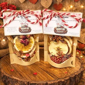May include: Two clear pouches filled with dried fruit and spices, tied with red and white twine. The pouches are labeled "Be My Valentine" and sit on a wooden slice. Decorative hearts and lights are in the background.