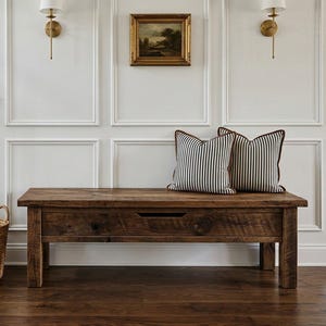 May include: A rustic wooden bench with a drawer, topped with two striped pillows, sits against a white paneled wall. A woven basket and a staircase with black railings and wooden steps are also visible.