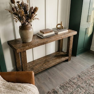 May include: A rustic wooden console table with a lower shelf, holding books and a gold decorative object. A large ceramic vase with dried flowers sits on the table. The room features a green cabinet, a leather chair, and a patterned rug.
