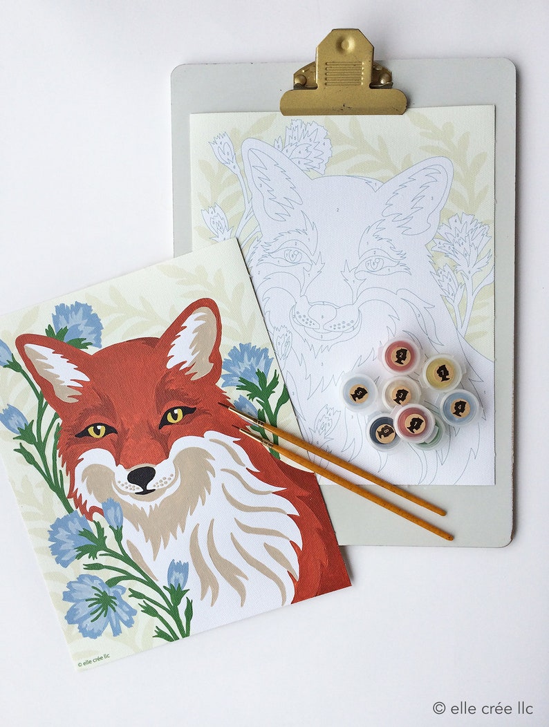Finished paint-by-number canvas beside unfinished canvas clipped to a clipboard. The design on both features a red fox surrounded by blue flowers. 8 acrylic paint pots are shown along with 2 paintbrushes that are included in each kit.