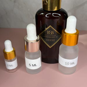 May include: A collection of brown glass bottles and frosted glass vials with gold and rose gold accents. The bottles and vials contain a nourishing body oil. The labels read "3 ML", "5 ML", and "10 ML". The large bottle has the text "BROWN SUGAR BABE NOURISHING BODY OIL MUSK BE NICE".