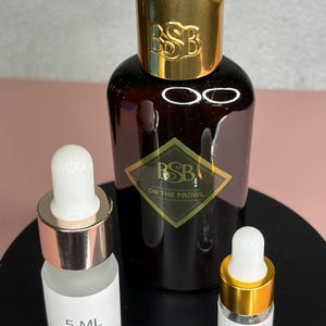 May include: A brown glass bottle with a gold cap and the letters "BSB" is flanked by two smaller bottles with droppers. One small bottle is labeled "5 ML" and the other "3 ML". The bottles are on a black surface.