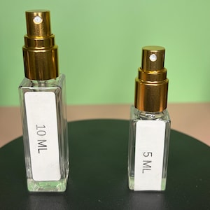 May include: Two clear glass perfume bottles with gold-colored spray tops. One bottle is labeled "10 ML" and the other is labeled "5 ML". The bottles are rectangular and sit on a dark surface against a green background.