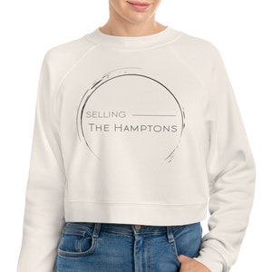 Könnte beinhalten: Weißer, kurzer Pullover mit einem schwarzen Kreis-Motiv und dem Text "SELLING THE HAMPTONS" in schwarzer und grauer Schrift.