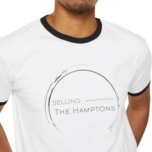 Puede incluir: Camiseta blanca con cuello negro y un gráfico de un círculo con el texto "SELLING THE HAMPTONS" dentro.