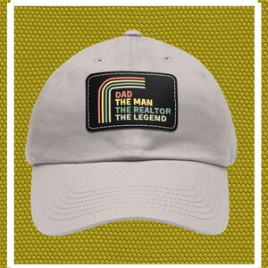 Op de afbeelding: Een grijze baseballpet met een zwart patch waarop "Dad The Man The Realtor The Legend" in een regenboogkleurig lettertype staat.