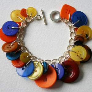 Puede incluir: Un brazalete de cadena de plata con una variedad de botones coloridos, incluyendo naranja, amarillo, azul y marrón. Los botones están unidos a la cadena con anillos de salto de plata.