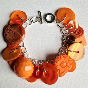 Peut inclure: Un bracelet à breloques en argent avec une variété de boutons orange et bruns. Les boutons sont de formes et de tailles différentes, et certains ont une finition brillante.