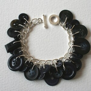 Può includere: Un braccialetto a catena d'argento con una chiusura a scorrimento e diversi bottoni neri attaccati. I bottoni sono di varie dimensioni e forme.