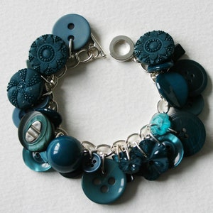 May include: Teal and turquoise button bracelet with a silver chain. The bracelet features a variety of buttons, including round, square, and flower-shaped buttons.