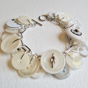 Könnte beinhalten: Ein handgefertigtes Armband mit weißen und cremefarbenen Knöpfen in verschiedenen Formen und Größen. Die Knöpfe sind durch silberfarbene Metallglieder und einen Knebelverschluss verbunden. Ein einzigartiges Accessoire.