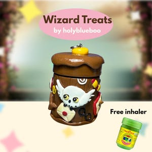 Op de afbeelding: Een bruine, cilindrische container met een deksel, versierd met een witte uil, een brief en andere kleurrijke accenten. De tekst "Wizard Treats" en "by holyblueboo" staan op een roze ovaal. Een kleine groene pot is ook zichtbaar.