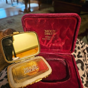May include: A gold compact with a red velvet case. The compact has a mirror on one side and a compartment for makeup on the other. The case is inscribed with the words "Moon Drops" and "Designed by Van Cleef & Arpels".