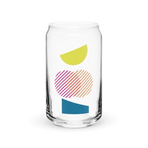 May include: A clear glass can with a geometric design. The design features a yellow crescent shape, a pink and orange striped oval, and a teal blue rectangle.