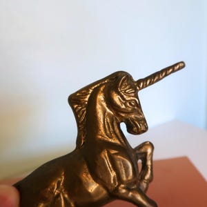 May include: A bronze-colored unicorn figurine. The mythical creature is posed with its front legs raised, and its horn is prominently displayed. The sculpture has detailed features and a polished finish, suggesting it is a decorative item.