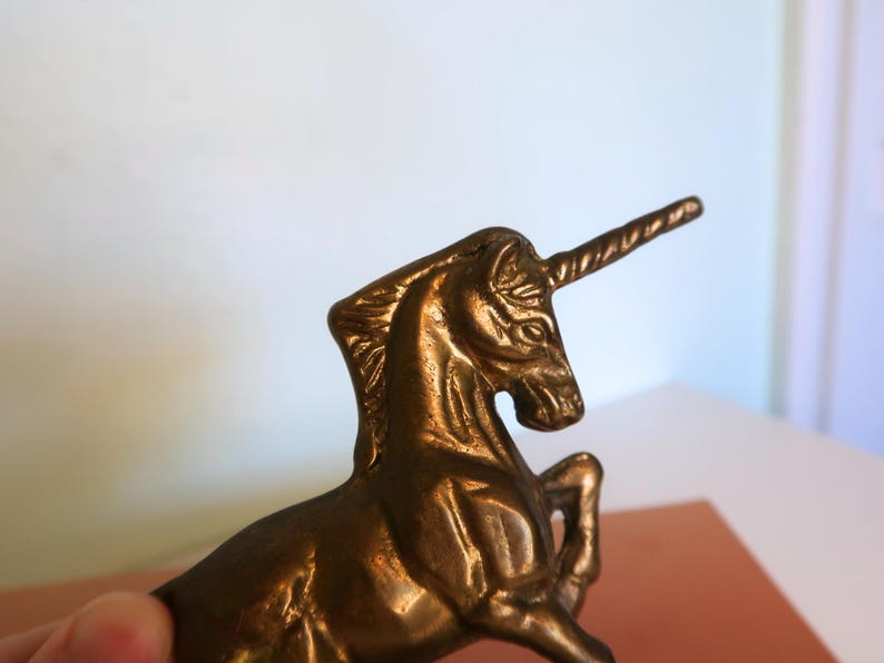 May include: A bronze-colored unicorn figurine. The mythical creature is posed with its front legs raised, and its horn is prominently displayed. The sculpture has detailed features and a polished finish, suggesting it is a decorative item.