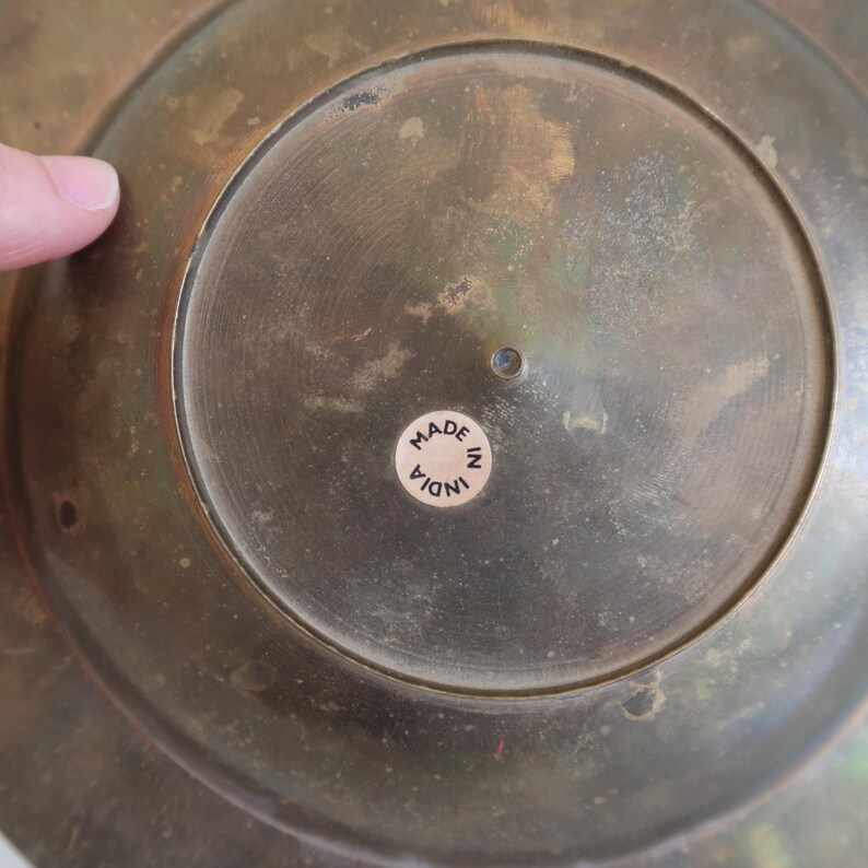 May include: Close-up of the underside of a brass bowl. The bowl has a circular design with a small sticker in the center that reads "MADE IN INDIA". The brass has a tarnished, aged appearance.