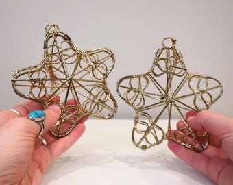 May include: Two gold-coloured, star-shaped ornaments. The ornaments are made of wire and have a delicate, open design. The stars are held up by hands. The background is a plain white surface.