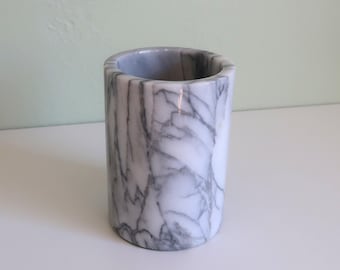 May include: A cylindrical marble container with a polished finish. The container is white with gray veining, and the interior is hollow. The object is likely a pencil holder or a small vase.