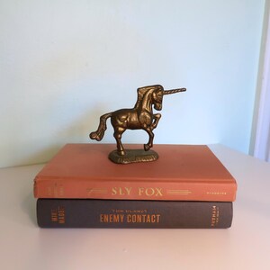 May include: A bronze unicorn figurine stands atop two vintage books. The top book is orange with the title "Sly Fox" in gold lettering. The bottom book is dark gray with the title "Enemy Contact".