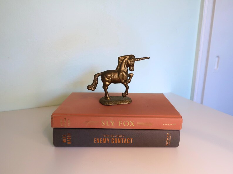May include: A bronze unicorn figurine stands atop two vintage books. The top book is orange with the title "Sly Fox" in gold lettering. The bottom book is dark gray with the title "Enemy Contact".