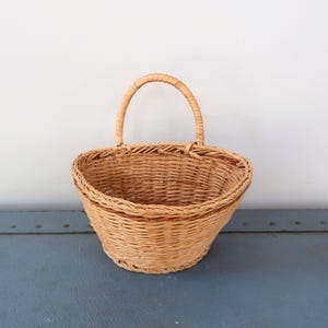 May include: A light brown, oval-shaped wicker basket with a curved handle. The basket has a woven design and a darker brown trim around the top edge. The handle is also made of woven wicker.