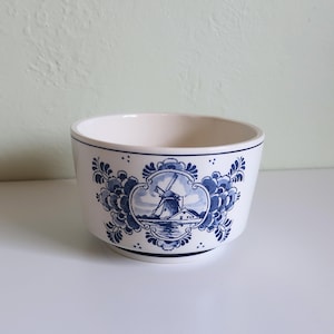Könnte beinhalten: Eine weiße Keramikschale mit blau-weißem Windmühlendesign. Die Schale hat eine abgerundete Form mit einem dunkelblauen Rand und einem dekorativen floralen Rand. Die Windmühlenszene ist von einem blauen Blumendesign umrahmt.