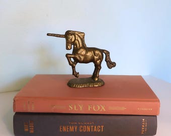 May include: A bronze unicorn figurine stands atop two books. The unicorn is detailed with a long horn and flowing mane. The books are a muted orange and dark brown, with gold lettering that reads "SLY FOX" and "ENEMY CONTACT."
