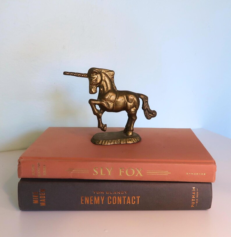 May include: A bronze unicorn figurine stands atop two books. The unicorn is detailed with a long horn and flowing mane. The books are a muted orange and dark brown, with gold lettering that reads "SLY FOX" and "ENEMY CONTACT."