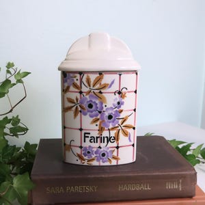 Puede incluir: Un bote de cerámica blanco con tapa, decorado con motivos florales morados y la palabra "Farine" en negro. El bote tiene un patrón de cuadrícula con líneas rosas y está sobre una pila de libros.