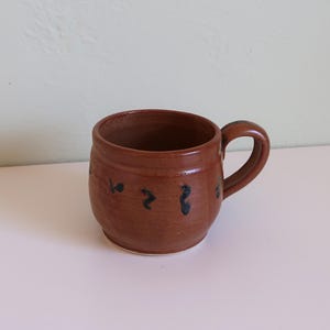 May include: A handmade brown ceramic mug with a rounded body and a curved handle. The mug features dark blue abstract markings around the body. The mug is suitable for coffee, tea, or other beverages.