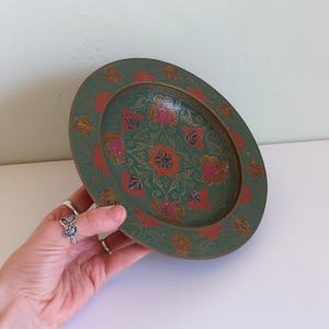 May include: An oval, green decorative tray with a floral design. The tray features a detailed pattern with red, pink, and gold accents. The tray is held by a hand.