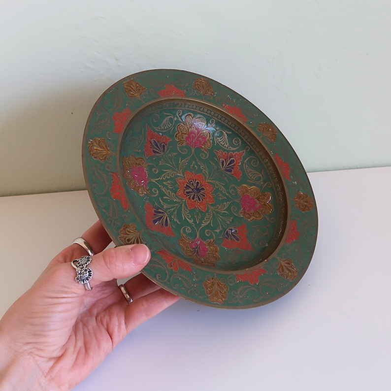 May include: An oval, green decorative tray with a floral design. The tray features a detailed pattern with red, pink, and gold accents. The tray is held by a hand.