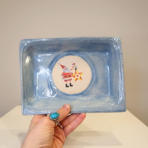 Puede incluir: Una bandeja rectangular de cerámica azul claro con una ilustración de Papá Noel en el centro. Papá Noel sostiene una linterna en forma de estrella. La bandeja tiene un borde texturizado y probablemente está hecha a mano.