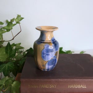 May include: A small ceramic vase with a beige top and a body decorated with blue floral patterns. The vase is sitting on a brown book with the title "Hardball" and the author's name "Sara Paretsky" visible.