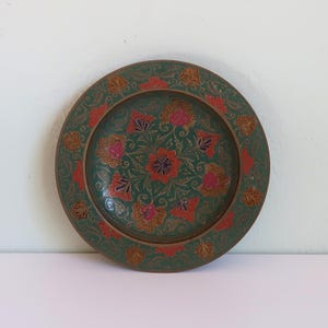 May include: A round, decorative plate with a green background and floral design. The plate features red, pink, and gold-colored flowers and leaves. The rim of the plate has a detailed, embossed pattern.