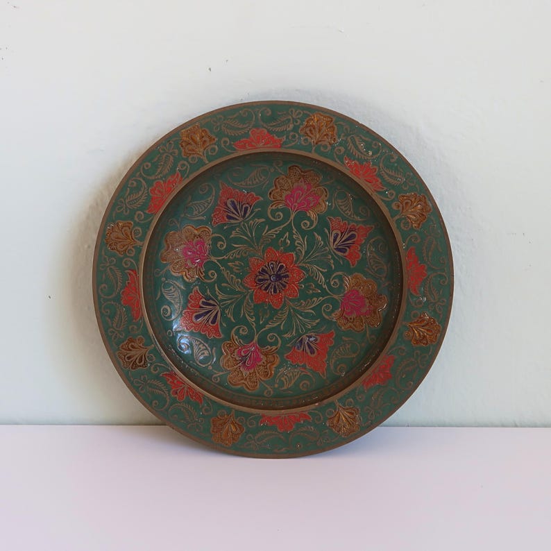 May include: A round, decorative plate with a green background and floral design. The plate features red, pink, and gold-colored flowers and leaves. The rim of the plate has a detailed, embossed pattern.