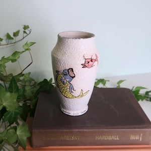 May include: A textured, off-white ceramic vase with hand-painted fish and animal illustrations. The vase features a wide mouth and a slightly tapered body. The fish is yellow and blue, and the animal is pink. The vase is sitting on a brown book.