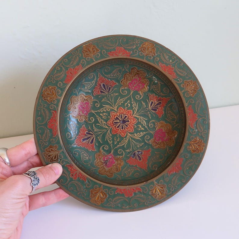 May include: A round, decorative plate with a teal background and floral design. The plate features a central flower with orange and gold details, surrounded by pink and red floral patterns. The rim has a gold border with additional floral accents.