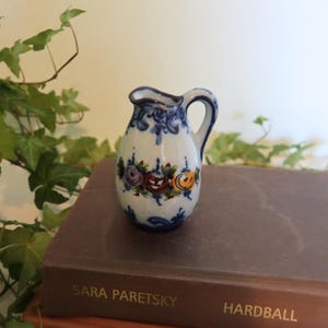 May include: A small, white ceramic pitcher with a blue handle and spout, decorated with blue floral patterns and colorful roses. The pitcher is resting on a brown book. The book's title is "Hardball".