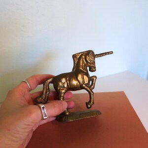 May include: A brass unicorn figurine with a detailed mane, tail, and horn. The mythical creature is standing on a rectangular base. The figurine is held in a hand, showcasing its size and intricate design. The unicorn is a golden color.