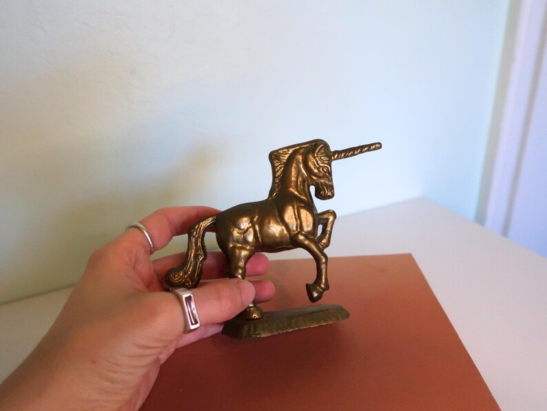 May include: A brass unicorn figurine with a detailed mane, tail, and horn. The mythical creature is standing on a rectangular base. The figurine is held in a hand, showcasing its size and intricate design. The unicorn is a golden color.